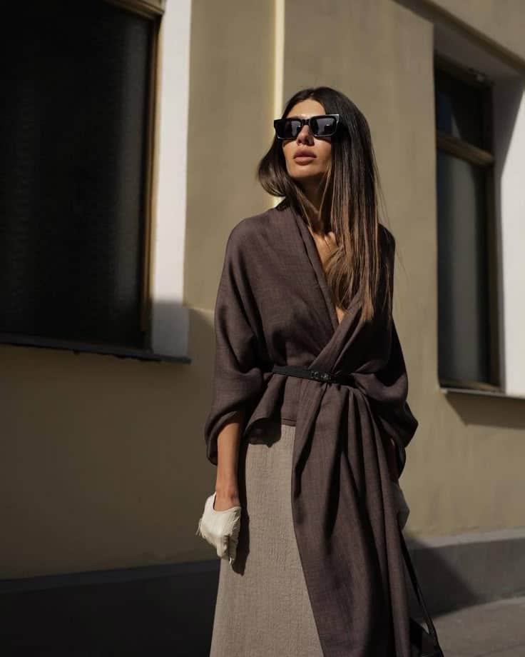 Stylish woman in sunglasses and brown wrap dress poses outdoors in sunlight.