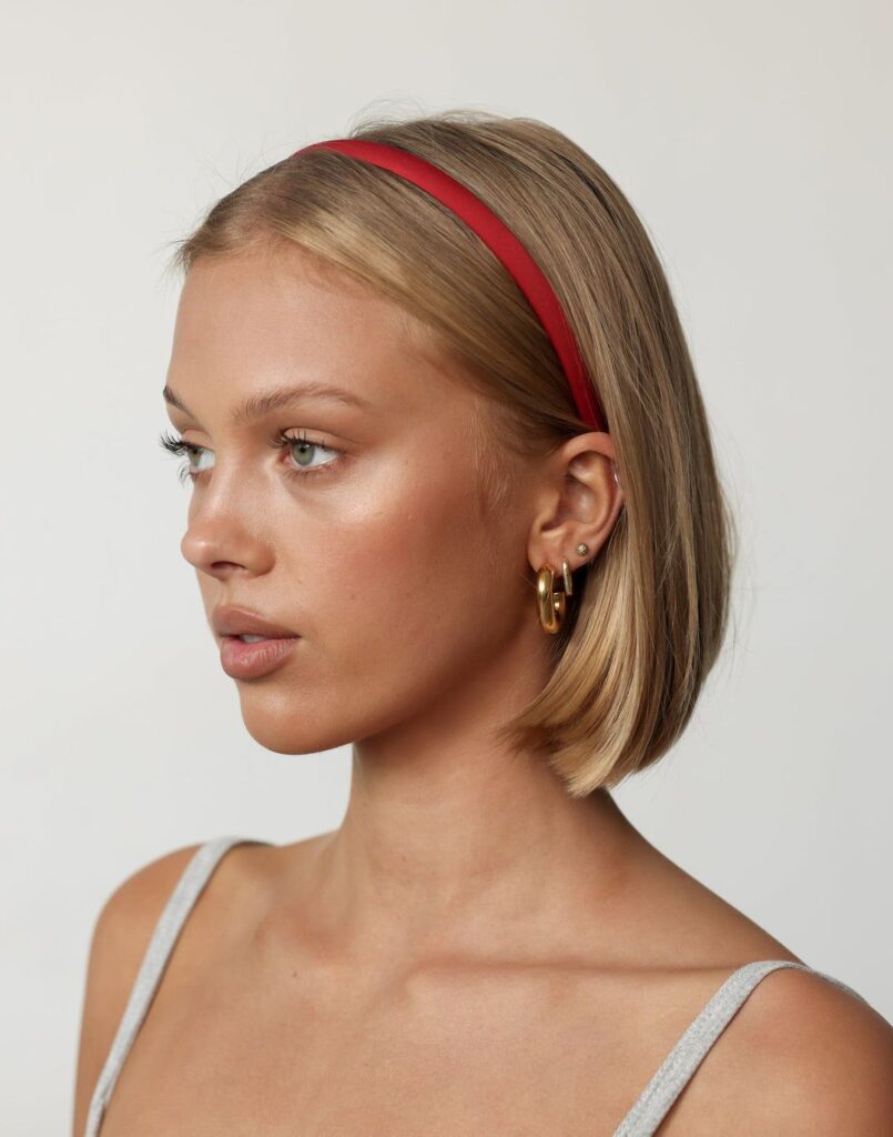 Woman with short blond hair wears a red headband and gold hoop earrings; side profile on white background.