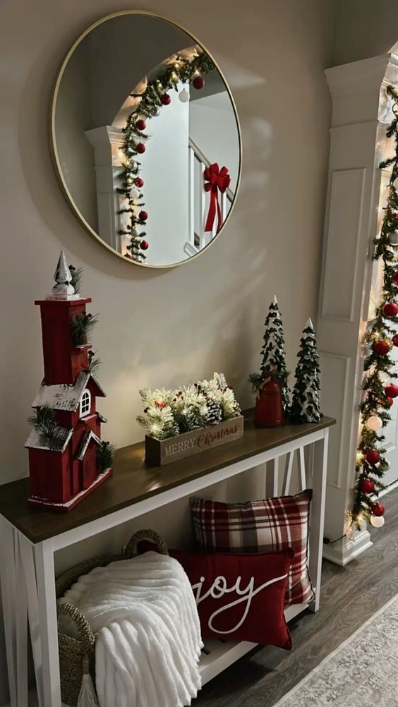 Festive holiday decor with a mirror, red accents, snowy houses, and greenery on a console table. Cozy Christmas scene.