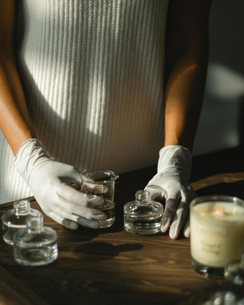 4a34347b699b7063c1d0a589d64e74e5 Person in white gloves handling glass beakers on a wooden table with a lit candle nearby. | Sky Rye Design Person in white gloves handling glass beakers on a wooden table with a lit candle nearby.