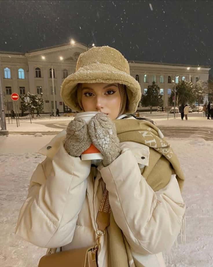 Person in winter attire holding a warm drink, standing in a snowy night cityscape with vintage buildings in the background.