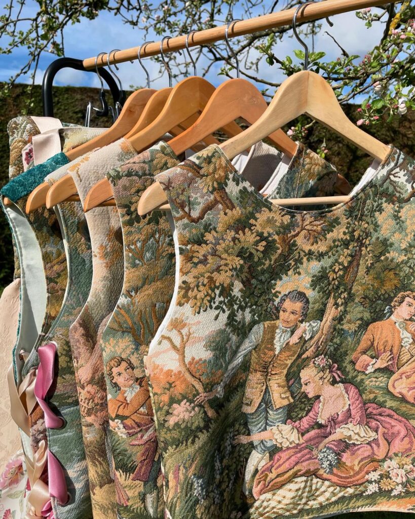 Vintage tapestry vests on hangers outdoors, featuring intricate historical scenes and vibrant colors.