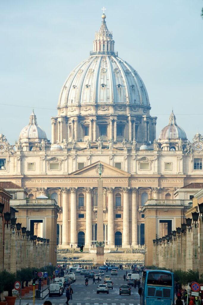 Majestic view of St. Peter's Basilica dome in Vatican City with busy street and vehicles in foreground.