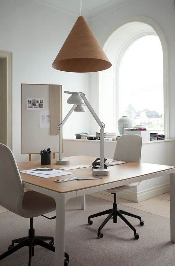 Modern office space with wooden desk, swivel chairs, and large lamp. Minimalist decor and natural light from arched window.