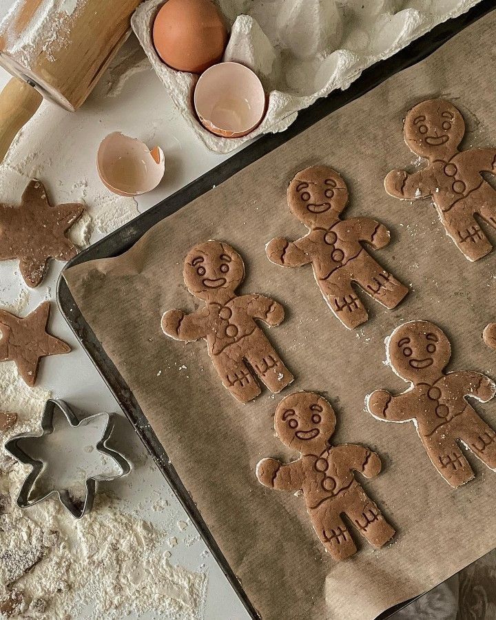 Gingerbread men cookies on a baking sheet with star cutters, eggshells, and flour scattered on a white surface.