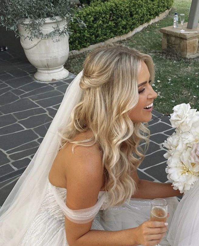 Smiling bride with blonde hair holding a bouquet and champagne, sitting on a tiled patio.