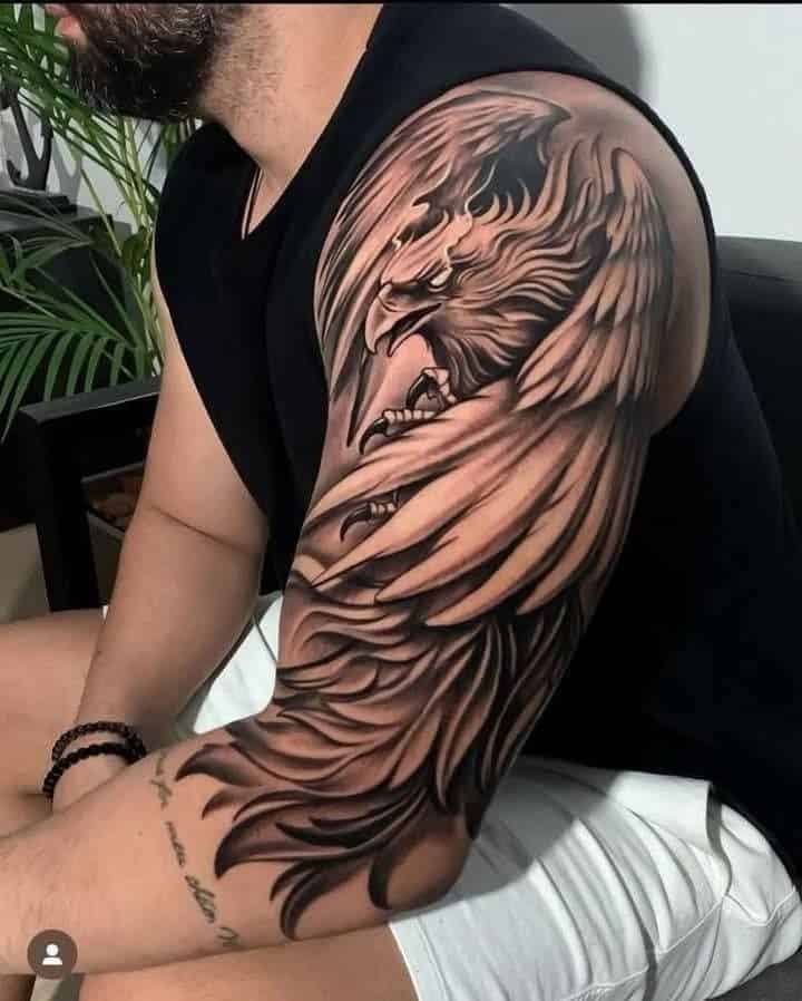 Man with detailed eagle tattoo on arm, wearing a black sleeveless shirt and white shorts, sitting indoors.