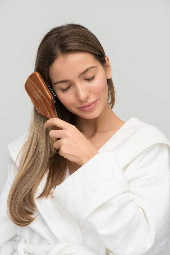 Woman in a white robe brushing her long hair, looking relaxed and enjoying self-care.