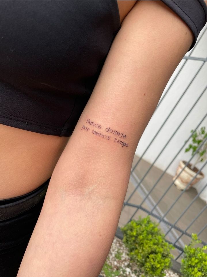Arm tattoo saying Nunca deseje por menos tempo on upper forearm, wearing a black top, background with plants.