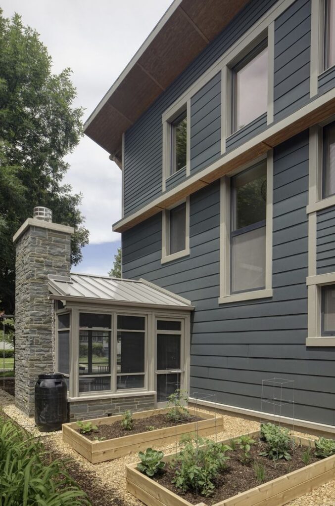 Modern house exterior with vegetable garden beds, metal roof, and stone chimney in daylight.