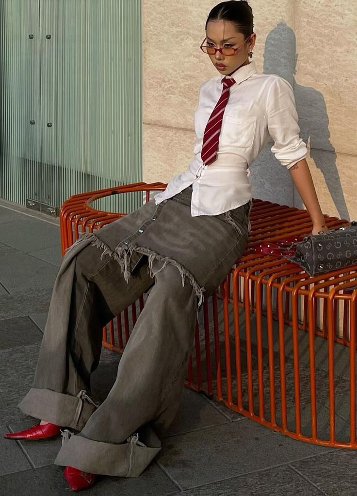 Fashion-forward person in white shirt, red tie, oversized jeans, and red heels sitting on an orange bench with confidence.