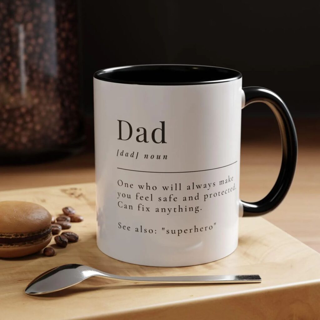 White mug with Dad definition, a macaron, and spoon on a wooden surface. Coffee beans scattered around.
