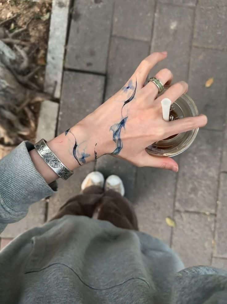 Hand with blue tattoo holding iced coffee in a plastic cup, wearing rings and a bracelet on a pavement background.