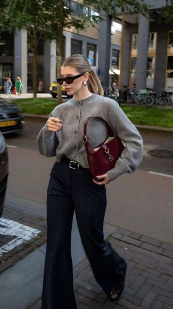 Stylish woman in the city wearing sunglasses, grey sweater, and pinstripe pants, carrying a burgundy handbag.