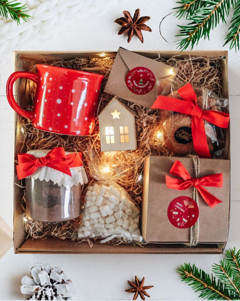 Holiday gift box with red mug, cocoa mix, marshmallows, lights, and wrapped presents on a festive background.