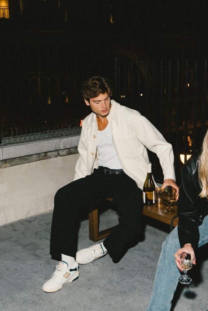 Man in white jacket and black pants sitting with a drink on a rooftop at night, enjoying a casual gathering.
