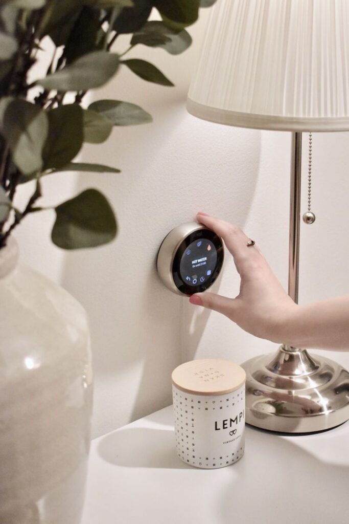 Hand adjusting smart thermostat on wall beside lamp and potted plant for home temperature control.