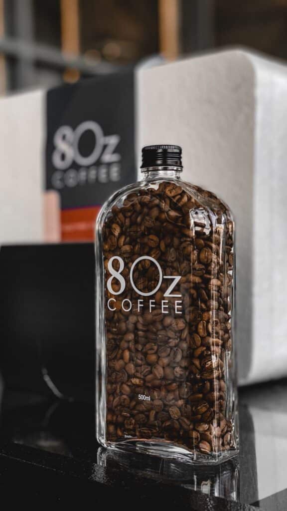 583686253a12d1deea6334dd16ce9a62 A stylish glass bottle filled with coffee beans, labeled 80z Coffee, placed on a reflective black surface. | Sky Rye Design A stylish glass bottle filled with coffee beans, labeled 80z Coffee, placed on a reflective black surface.