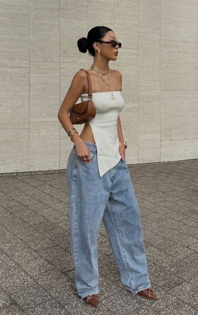 Woman in a trendy outfit with oversized jeans, white top, and brown accessories, posing stylishly outdoors.