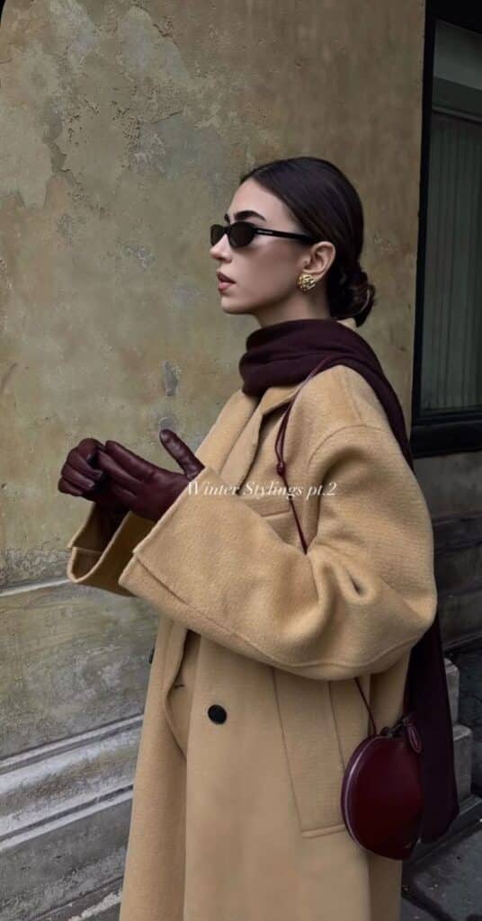 Fashionable woman in sunglasses wearing a stylish coat, scarf, gloves, and crossbody bag for winter styling.