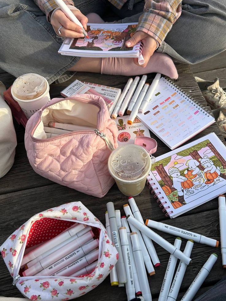 Person coloring in sketchbook outdoors, surrounded by markers, notebooks, and coffee, creating a cozy art scene.