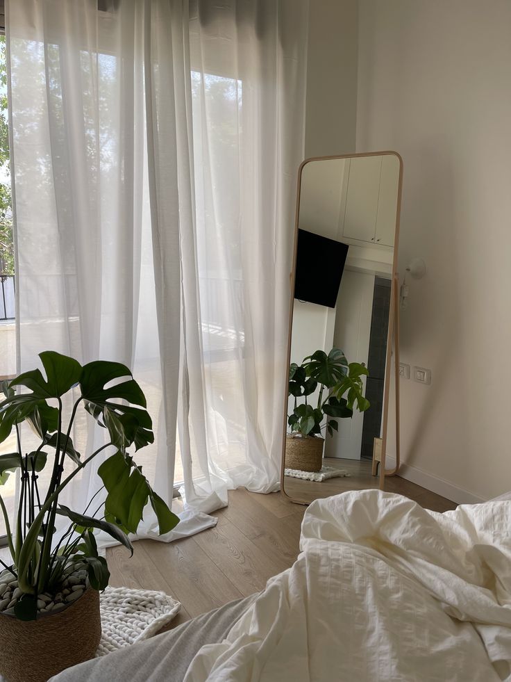 5c00146c-af8f-41e8-84a8-b9661a06324b Bright bedroom with large mirror, potted plant, and sheer curtains allowing soft sunlight. Cozy and natural ambiance. | Sky Rye Design Bright bedroom with large mirror, potted plant, and sheer curtains allowing soft sunlight. Cozy and natural ambiance.