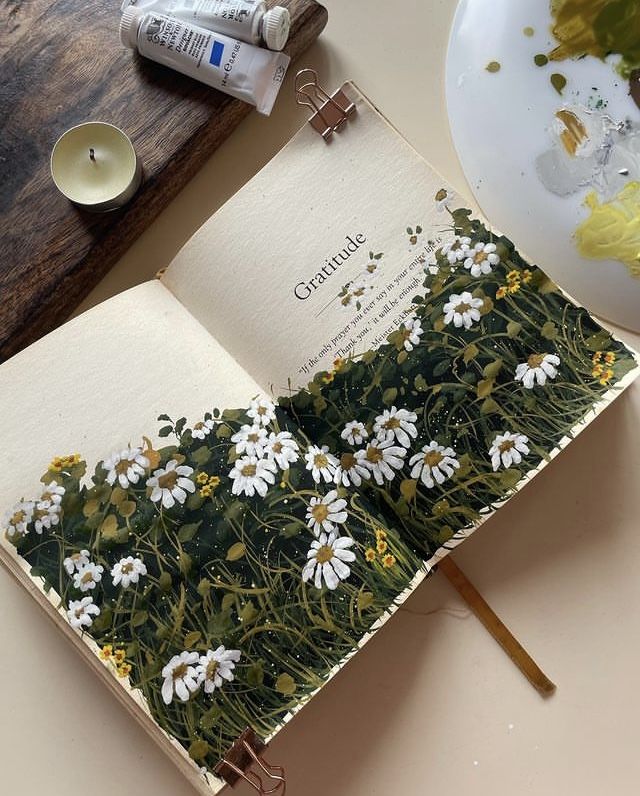 Open book with painted daisies and Gratitude text, next to candle and paint tubes on a table.