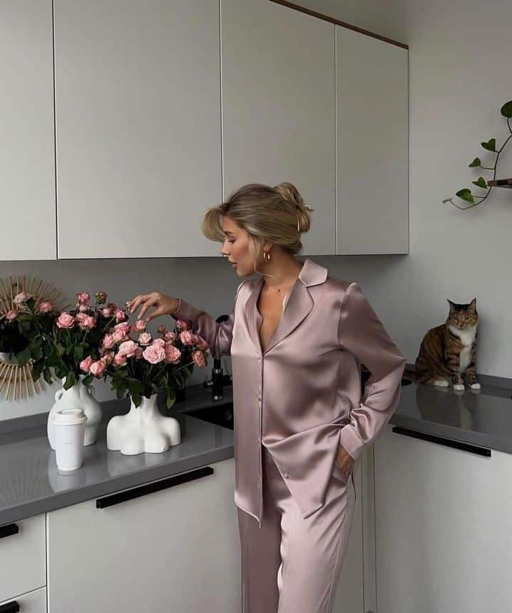 Woman in satin pajamas admires pink roses in modern kitchen, with a cat sitting on the counter.
