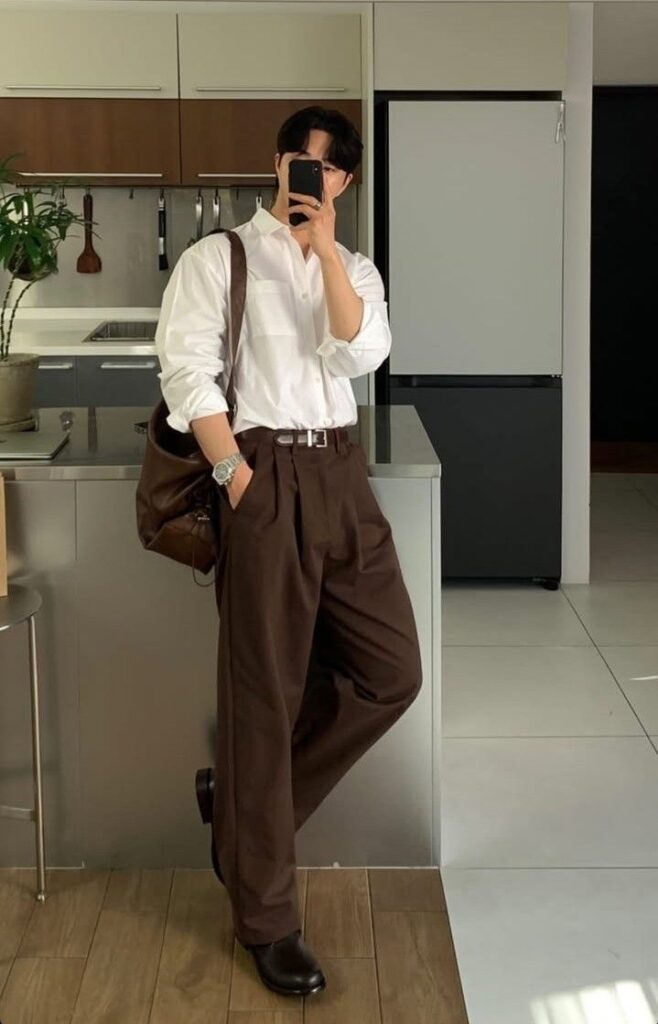 Man in smart casual outfit posing with phone in modern kitchen, wearing white shirt and brown trousers.