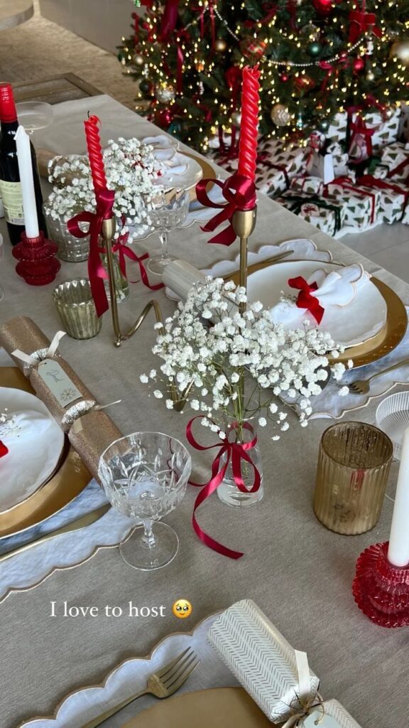 Festive holiday table setting with red candles, white flowers, and a decorated Christmas tree in the background.