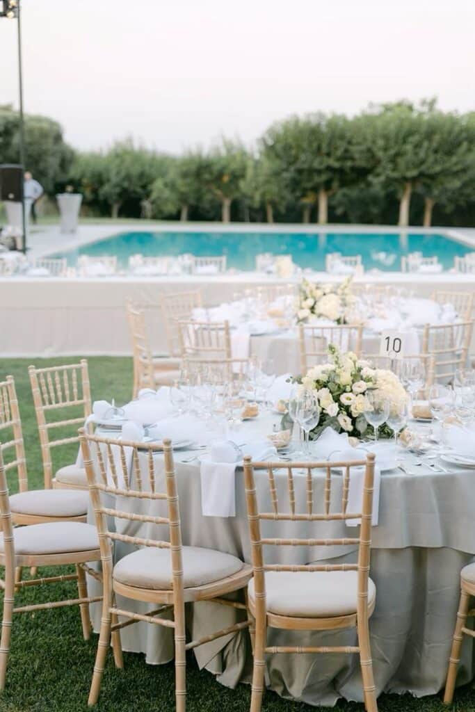Elegant outdoor wedding reception with tables, floral centerpieces, and poolside view against a backdrop of trees.