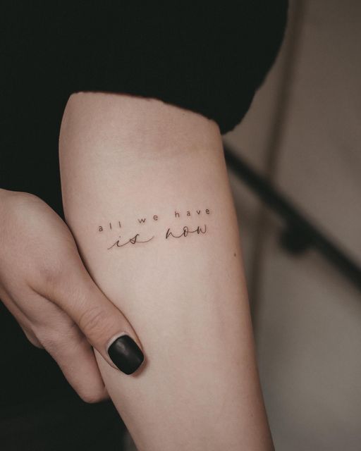 Minimalist tattoo on arm with the phrase all we have is now, held by a hand with black nail polish.