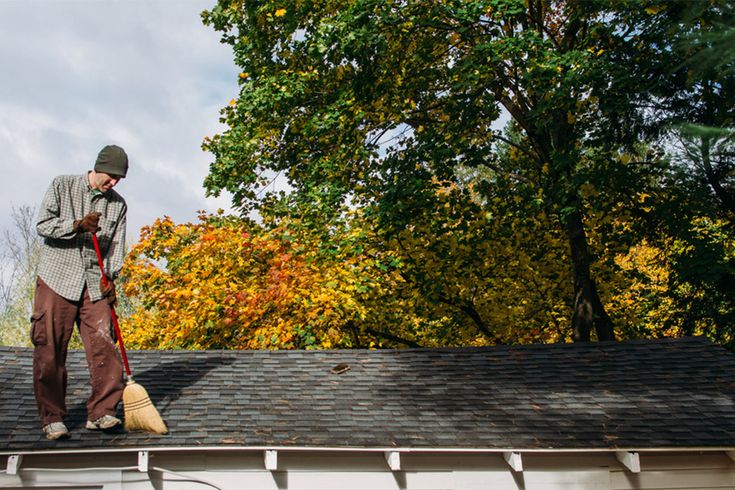 Man cleaning leaves off roof with broom on a sunny autumn day, surrounded by colorful trees.