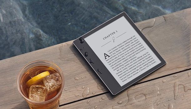 E-reader with an open book next to a refreshing iced tea on a wooden deck by the water.
