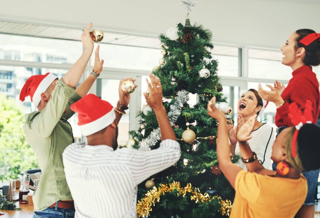 Office team decorating Christmas tree with ornaments and Santa hats, celebrating the holiday season indoors.