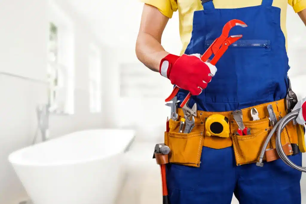 Plumber in blue overalls holding wrench, tool belt full, standing in bright bathroom near bathtub.