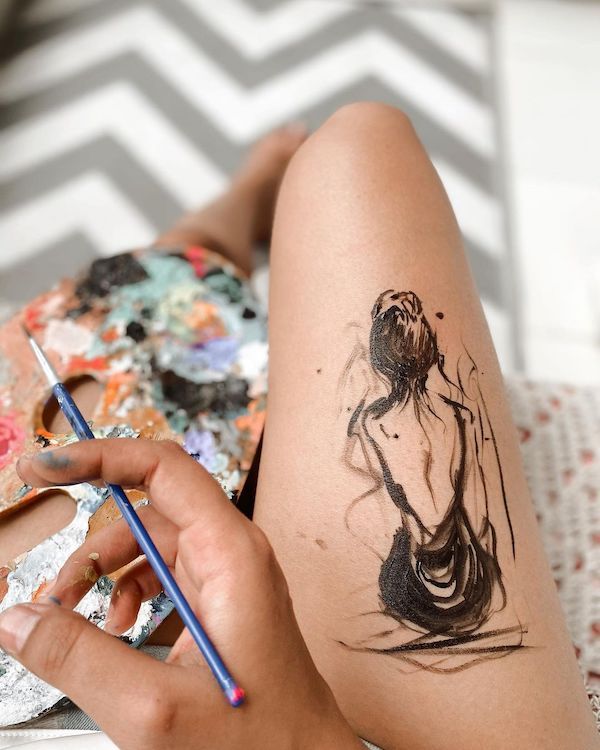 66 Alluring Thigh Tattoos For Women With Meaning - Our Mindful Life Hand painting an abstract figure on the thigh with a paintbrush and colorful palette, on a zigzag patterned background. | Sky Rye Design Hand painting an abstract figure on the thigh with a paintbrush and colorful palette, on a zigzag patterned background.