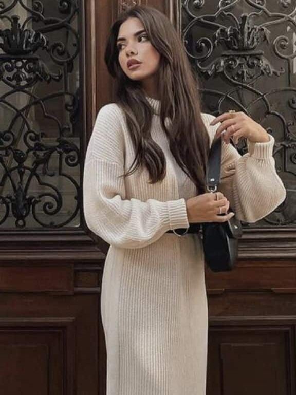 Woman in a cream knitted dress with white sneakers, holding a black bag in front of an ornate door.