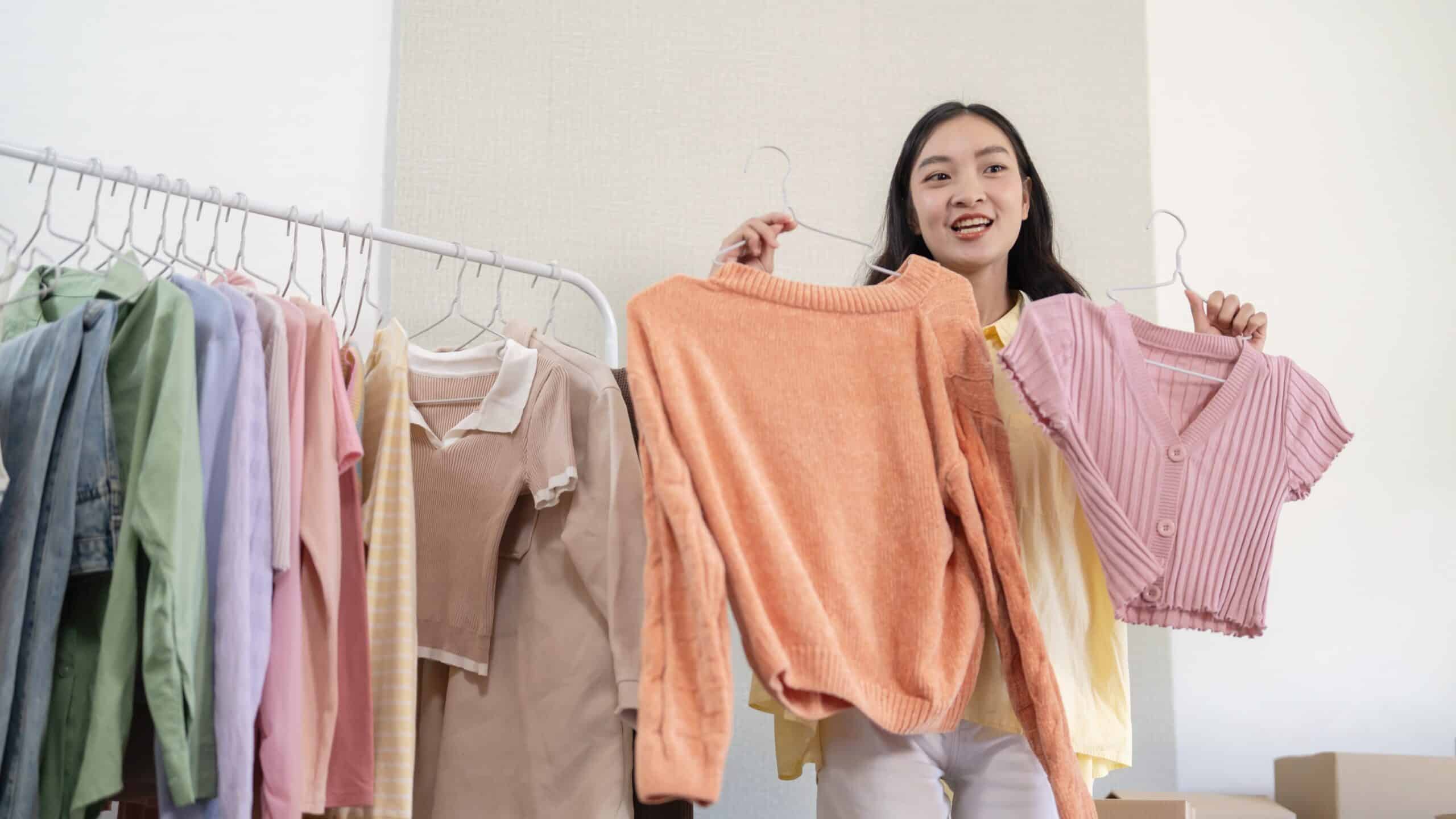 67c19db71f63a229af7eee01_cropped-shutterstock_2390502287 Smiling woman showcasing orange sweater and pink top, surrounded by colorful hanging clothes. | Sky Rye Design Smiling woman showcasing orange sweater and pink top, surrounded by colorful hanging clothes.