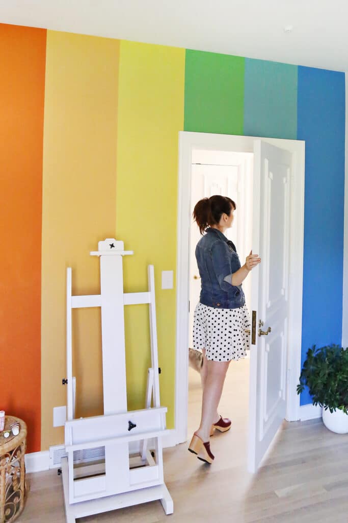 Woman in polka dot dress entering room with rainbow-striped walls, next to a white easel and a green plant.