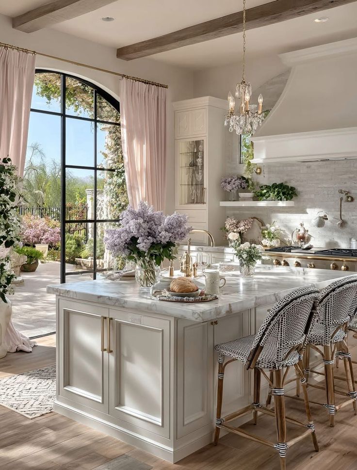 Luxurious kitchen with marble island, elegant chandelier, and garden view through large windows.