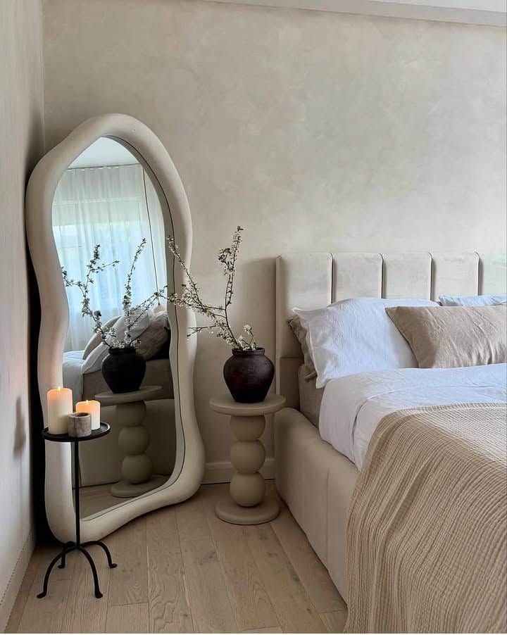Modern bedroom with wavy mirror, stylish side table, candles, and neutral bedding for a cozy and elegant atmosphere.