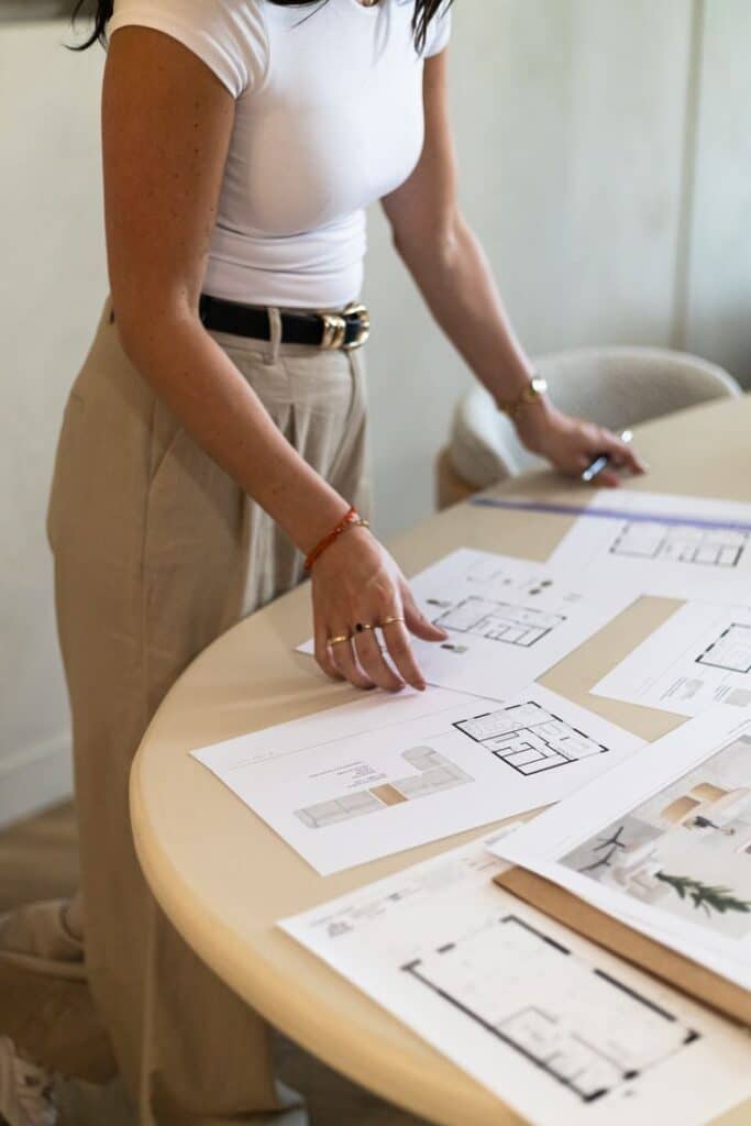 6bfe1e1b6b59e994702a27016e41a6c4 Person reviewing architectural floor plans on a table, wearing a white shirt and beige pants. | Sky Rye Design Person reviewing architectural floor plans on a table, wearing a white shirt and beige pants.