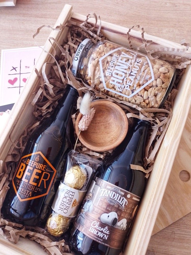 Gift box with craft beer, honey peanuts, Ferrero Rocher, and wooden bowl on a rustic wooden surface.