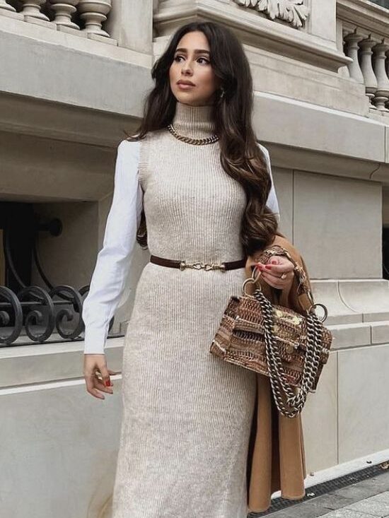 Stylish woman in sleeveless knit dress, brown boots, and carrying a patterned handbag on a city street.