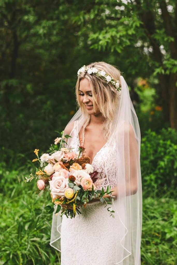 Bride in lace gown with floral bouquet and crown, looking down in a lush green garden setting.