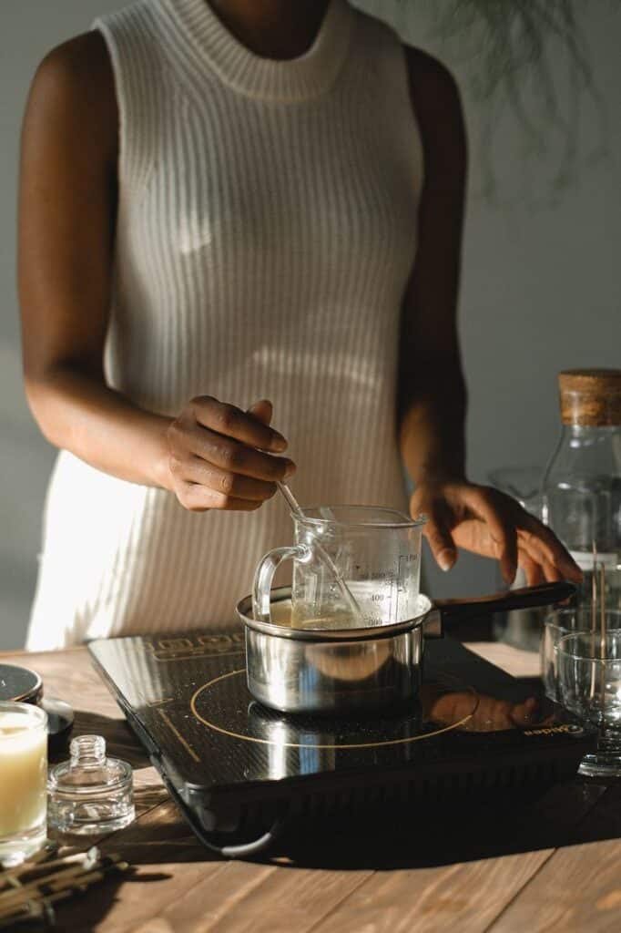 _ 7 Person in a white sweater melting wax on an induction stove, stirring with a silver spoon in a glass beaker. | Sky Rye Design Person in a white sweater melting wax on an induction stove, stirring with a silver spoon in a glass beaker.