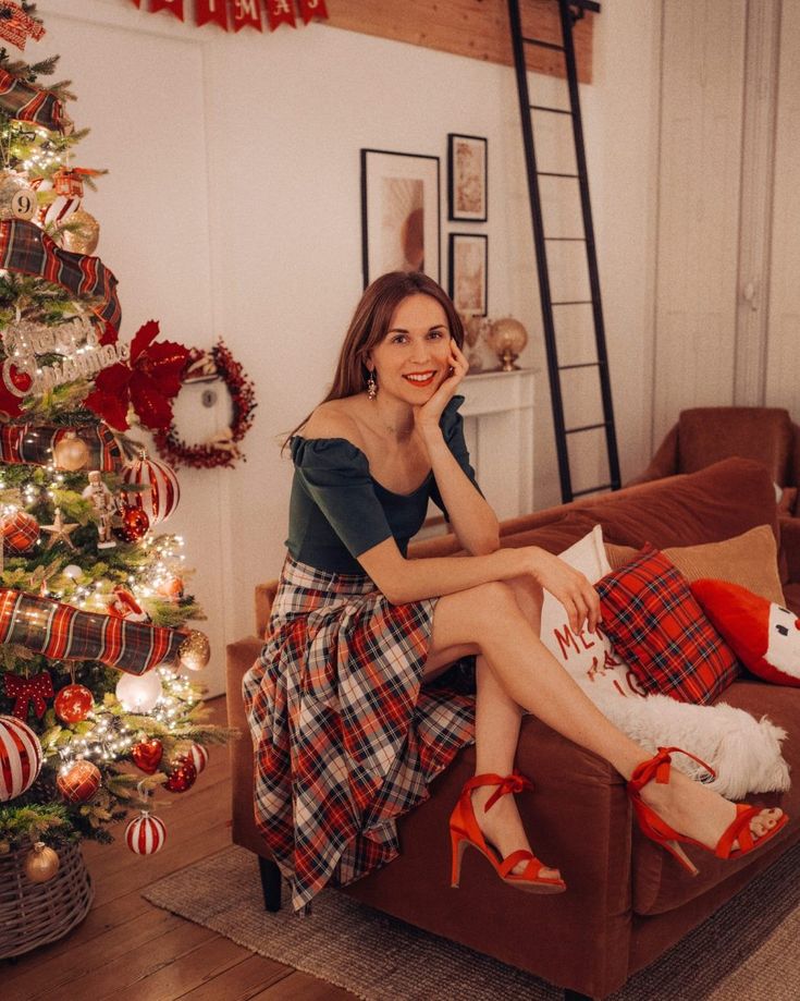Woman in festive attire sitting by a Christmas tree in a cozy living room.