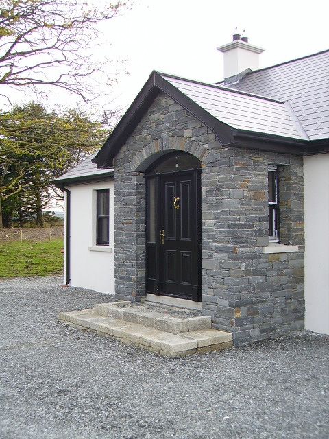 _ 7 Stone house entrance with black door, arched stonework, and steps, surrounded by gravel, trees in the background. | Sky Rye Design Stone house entrance with black door, arched stonework, and steps, surrounded by gravel, trees in the background.