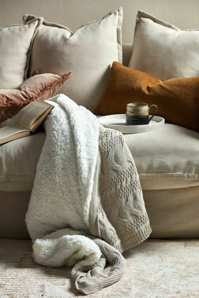 _ 7 Cozy beige sofa with plush pillows, warm knit blanket, open book, and coffee cup on tray. Perfect for relaxation. | Sky Rye Design Cozy beige sofa with plush pillows, warm knit blanket, open book, and coffee cup on tray. Perfect for relaxation.